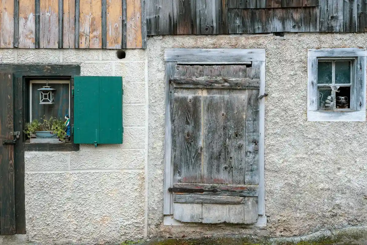 Exterior of an old building showing a weathered wooden door, a small window with a green shutter, and rough textured walls with weathered wood above.