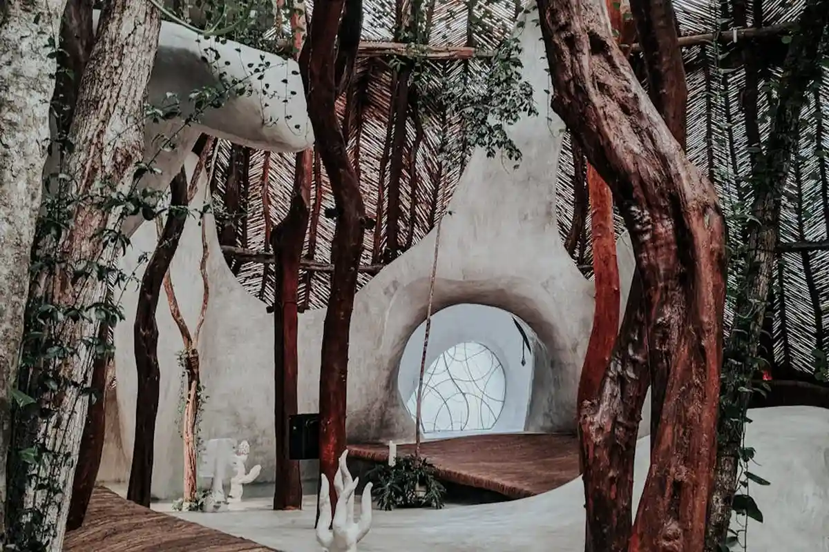 Interior scene with sculptural tree-like trunks, curved white walls, and a circular window, blending natural forms with architectural space.