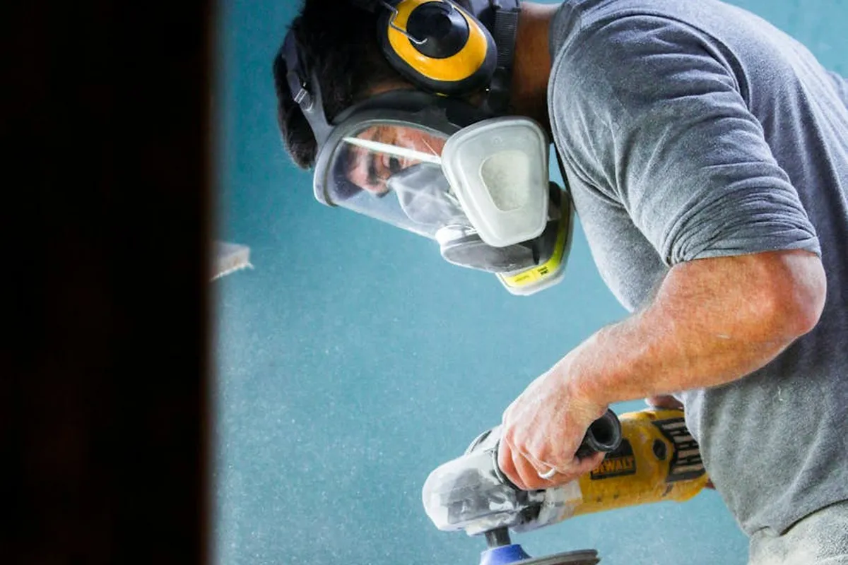Worker wearing a respirator mask, protective eyewear, and hearing protection while operating a power drill in a woodworking workshop.