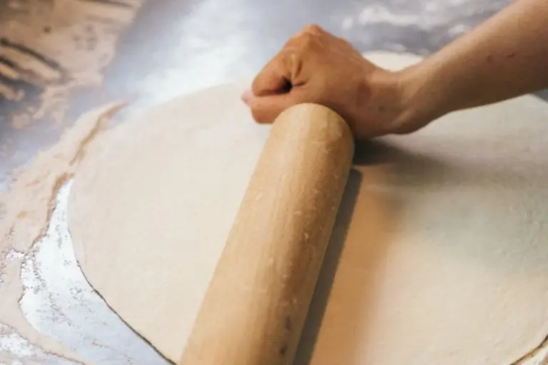Close-up of a wooden rolling pin being rolled over dough on a floured surface.
