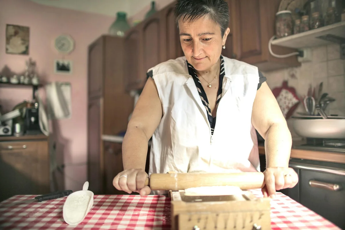 A person in a home kitchen uses a wooden rolling pin to flatten dough on a red-and-white checkered tablecloth.