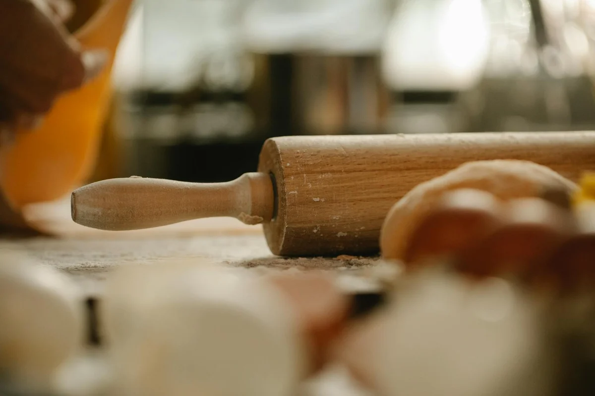 A wooden rolling pin rests on a kitchen counter with dough and eggs blurred in the foreground.