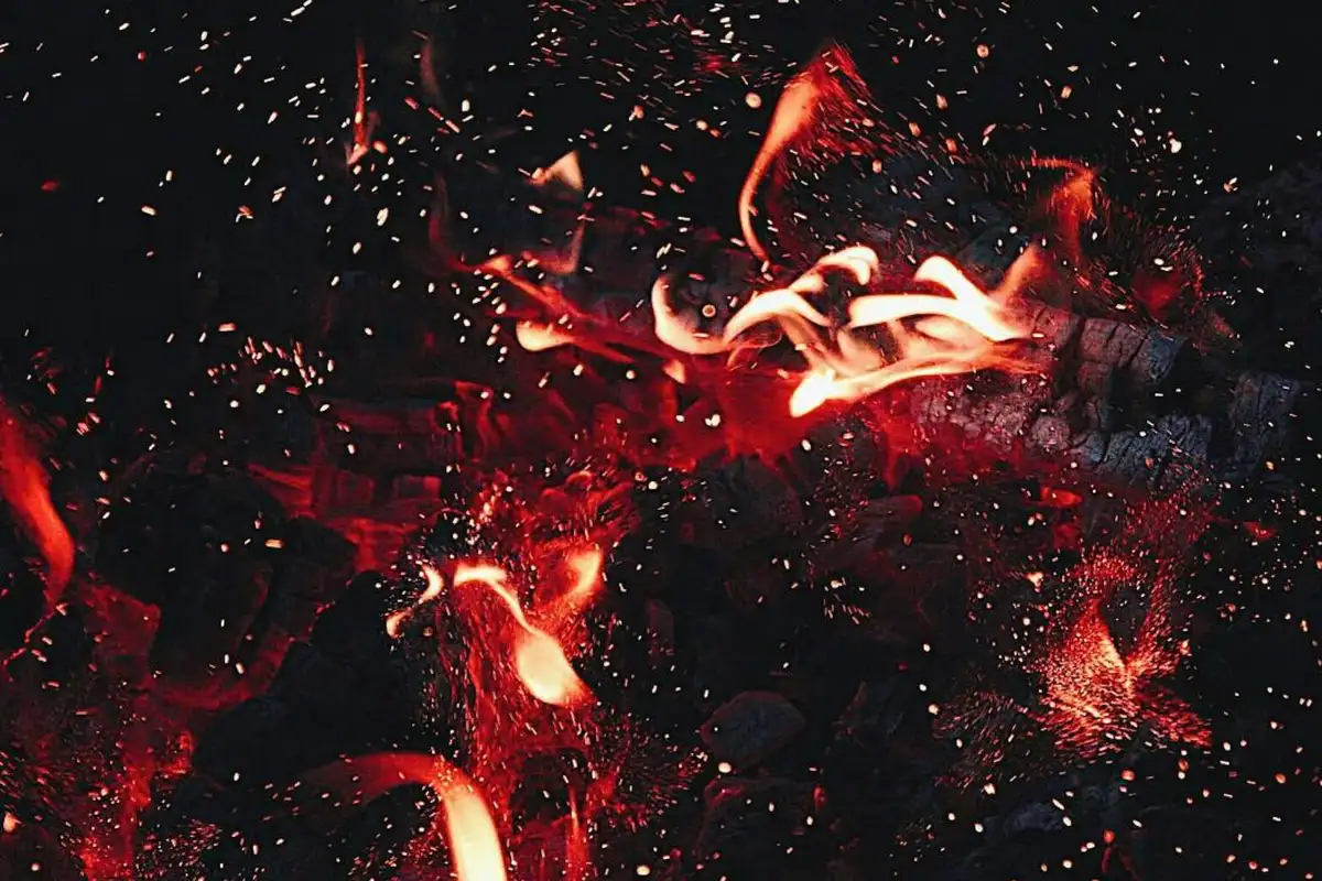 Close-up of glowing embers and flickering flames in a wood fire with small sparks rising.