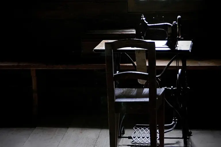 Dimly lit workshop with an antique sewing machine on a table and a wooden chair in the foreground.