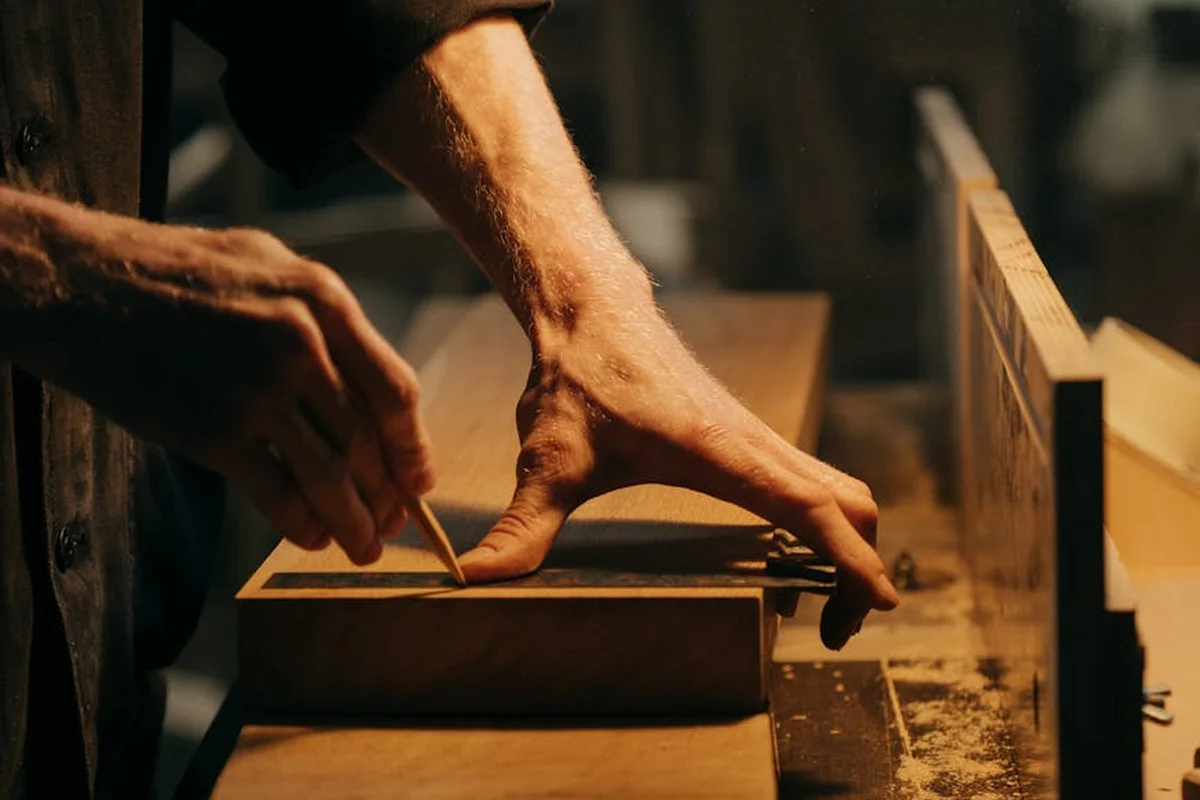 A woodworker's hands guide a chisel along a wooden board in a dimly lit workshop, with sawdust on the workbench.