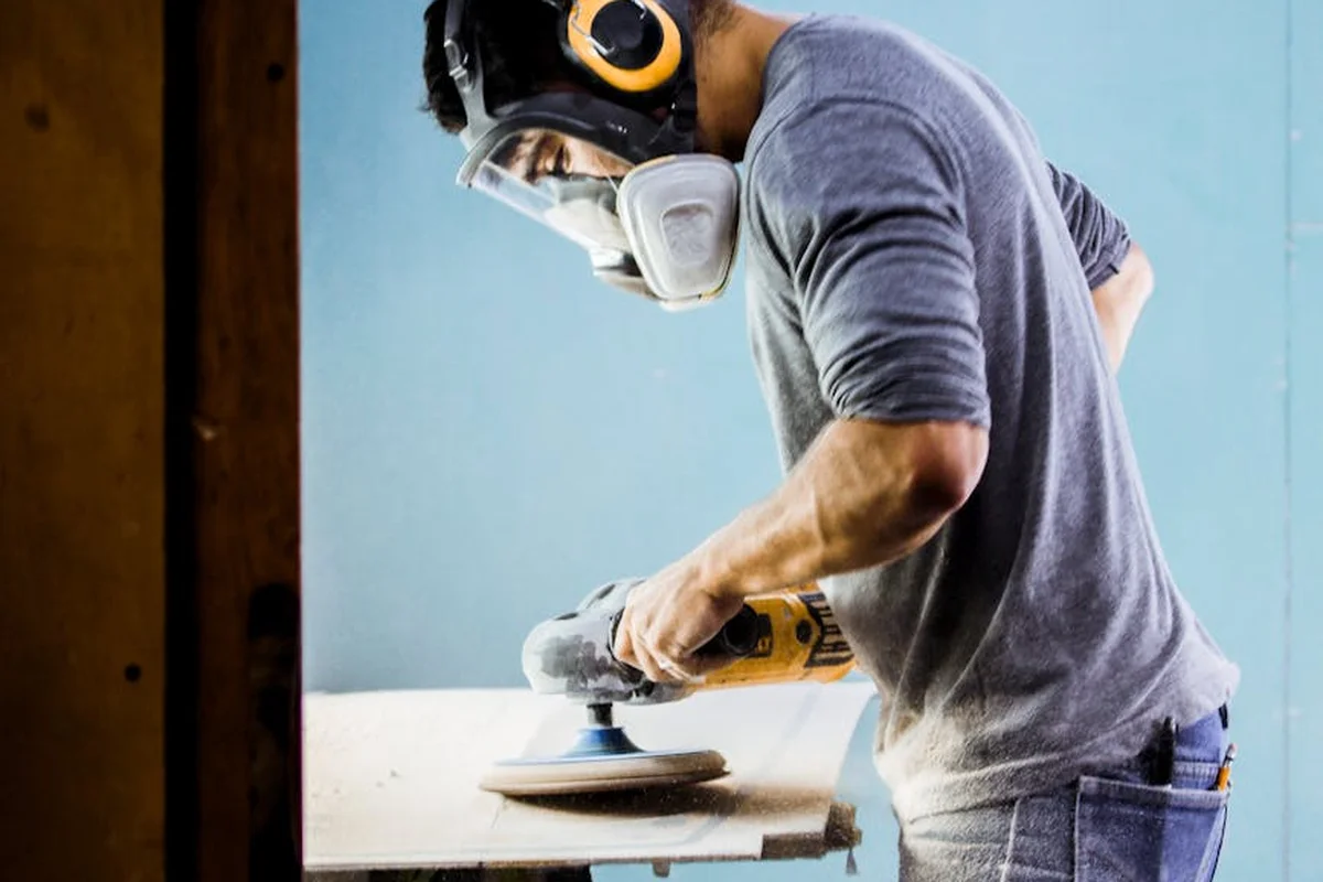 Person wearing a respirator and safety goggles using a palm sander to sand a wooden surface