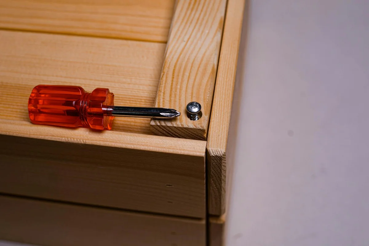 A screwdriver with an orange handle sits on a light wooden surface beside a small metal screw, illustrating the initial stage of removing a stubborn wood screw.