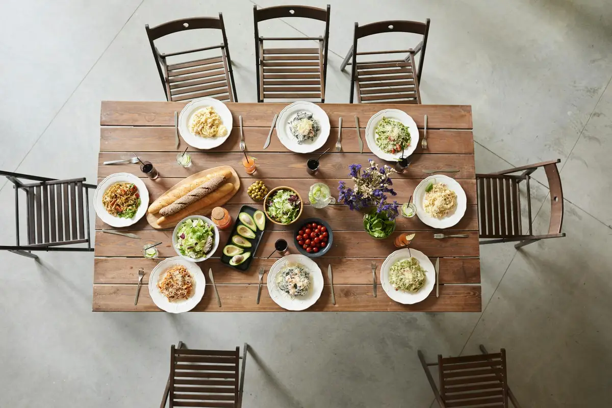 Top-down view of a rustic wooden dining table set for a meal with eight chairs around it, plates of salads and other dishes, and a central bouquet, highlighting natural wood tones.