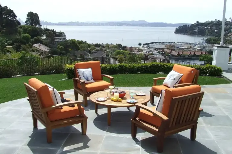 Teak outdoor furniture set with orange cushions arranged around a round coffee table on a stone patio, with a harbor view in the background.