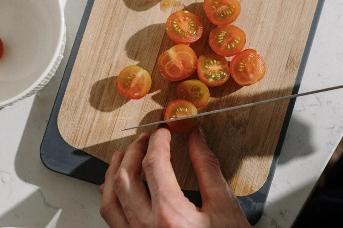 Close-up of a hand slicing cherry tomatoes on a teak cutting board placed on a kitchen counter, with a knife resting on the board.