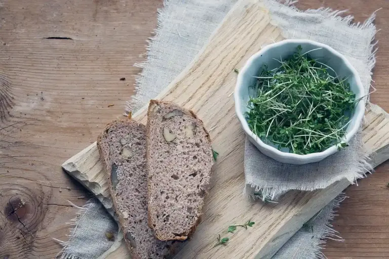 A wooden cutting board on a rustic wooden table with slices of brown bread and a small white bowl of fresh greens.