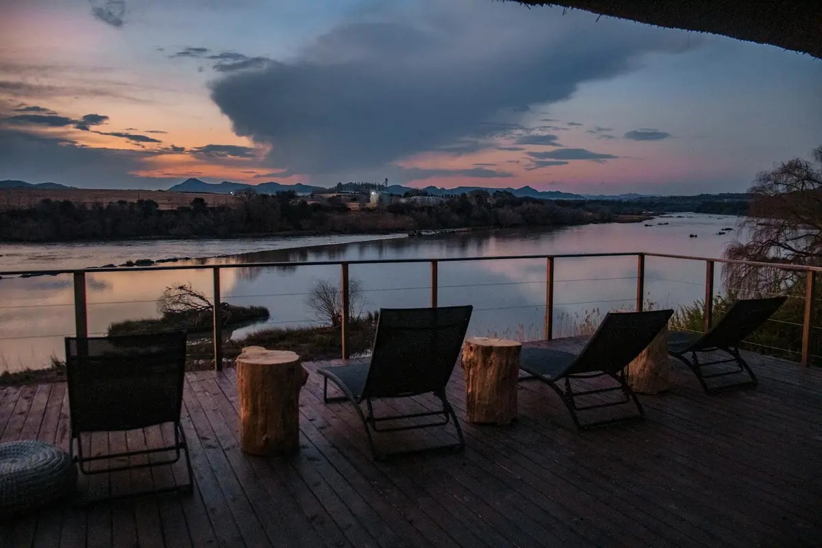 Sunset over a teak deck with lounge chairs and stump-like side tables overlooking a calm river.