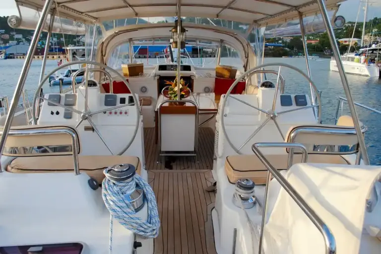 View of a luxury sailing yacht's teak deck with two helm wheels, ropes, and a marina in the background.