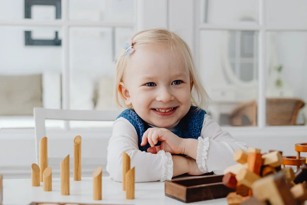 Smiling toddler seated at a table with wooden building blocks and toys in a bright, tidy playroom