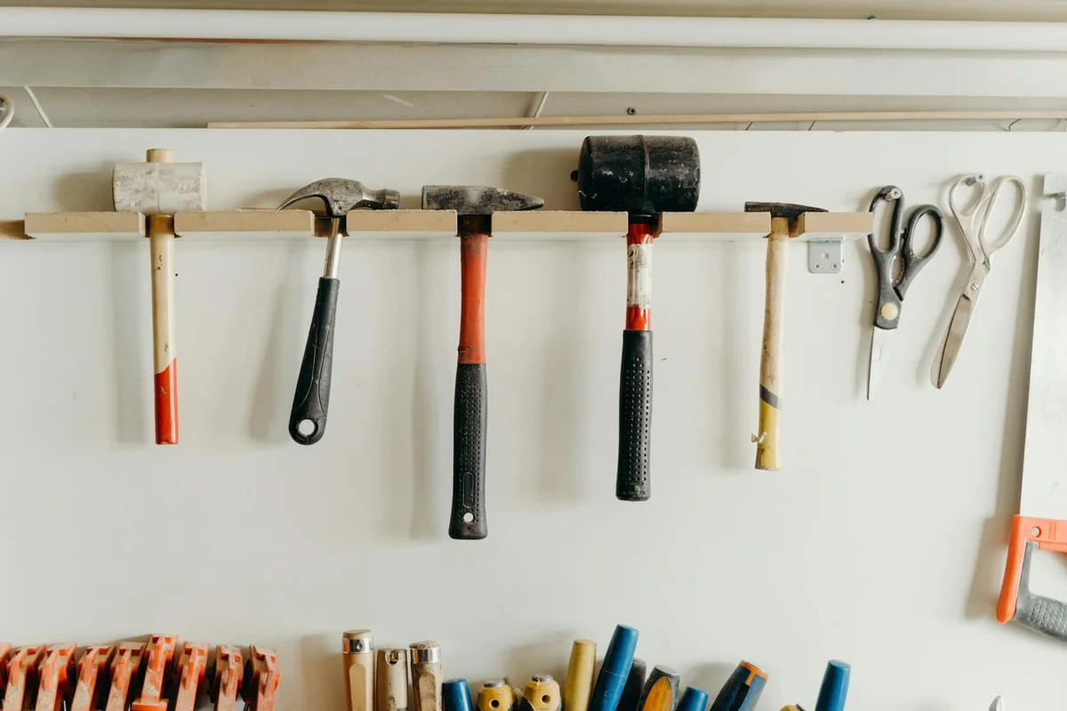 Row of hand tools hung on a wall in a woodworking shop, including hammers, mallets, chisels, and scissors, ready for joinery tasks.