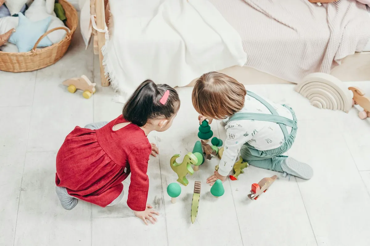 Two children kneeling on a light wooden floor, playing with wooden toys and shapes in a bright playroom.