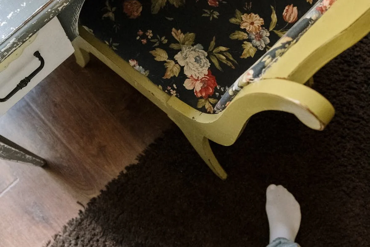 Close-up of a yellow-painted wooden chair with a curved leg and floral upholstered seat, on a dark rug, with a white sock visible at the bottom edge.