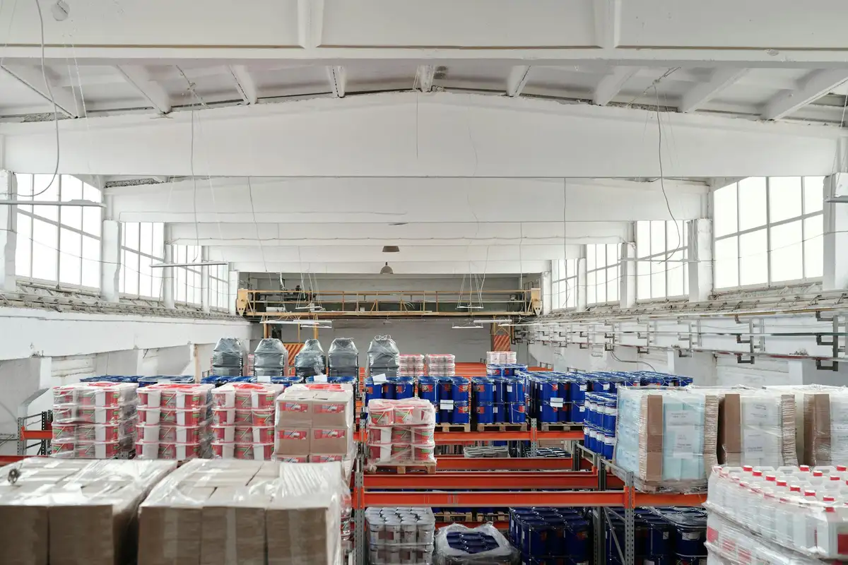 Interior of a large warehouse with multiple rows of pallets wrapped in plastic, organized on pallets and shelves under bright lighting.