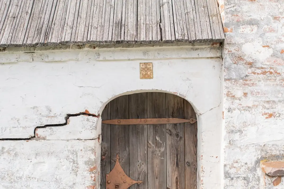 Weathered wooden door set in a cracked white plaster wall with an arched doorway and exposed hinge; the door appears warped.