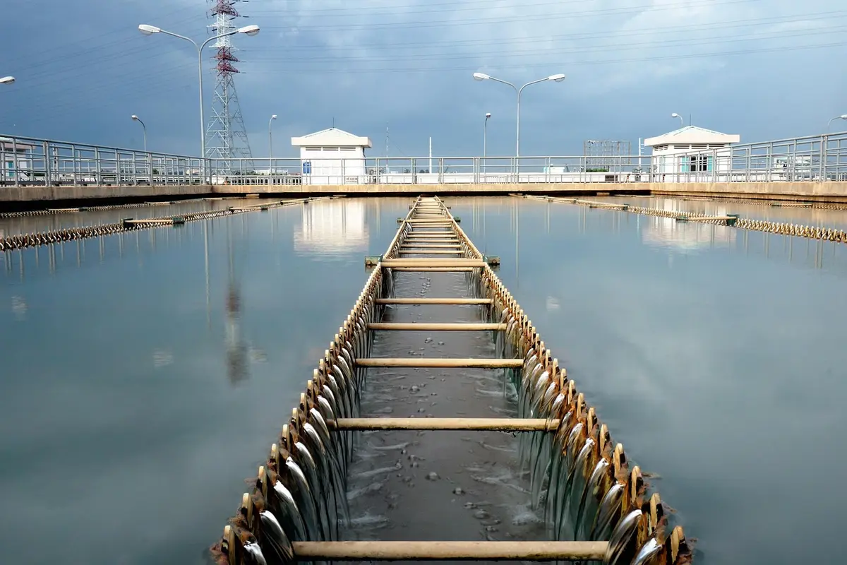 Outdoor water treatment facility with a narrow wooden frame extending into a large pool, flanked by metal walkways and control buildings in the distance.