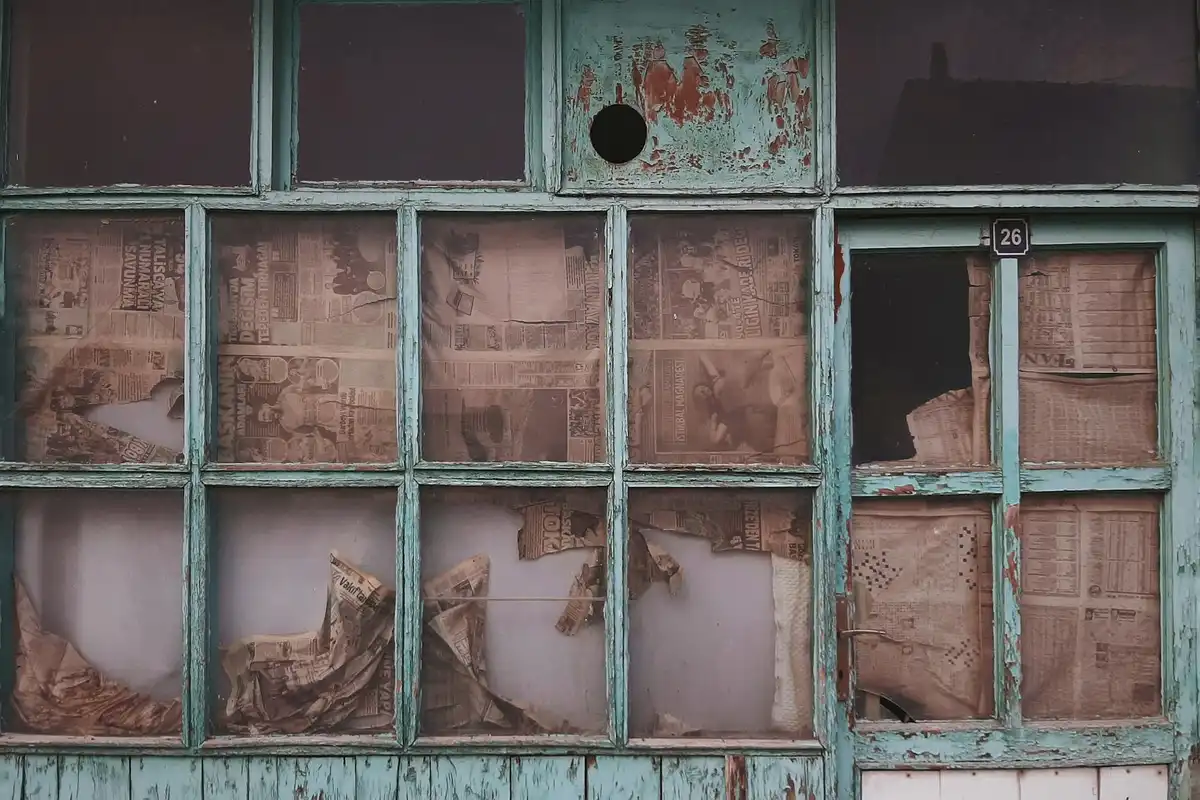 Weathered greenish-teal window frame with rotted wood and torn newspaper behind the panes, illustrating signs of structural wood damage.