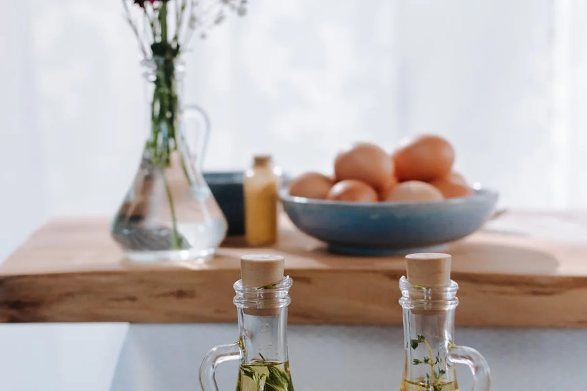 A wooden kitchen countertop with oil bottles in the foreground, a cutting board and a bowl of eggs in the background, and a vase with flowers.