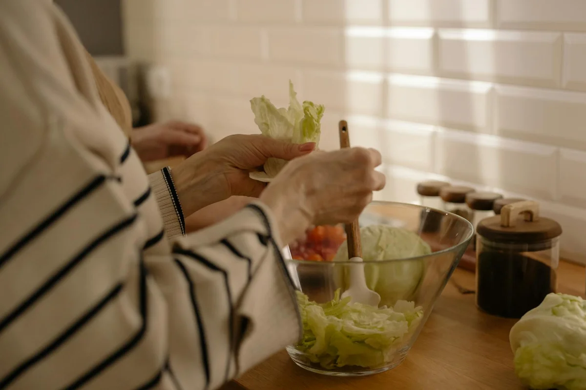 Person at a wooden kitchen counter washing lettuce in a glass mixing bowl; jars and kitchen items in the background