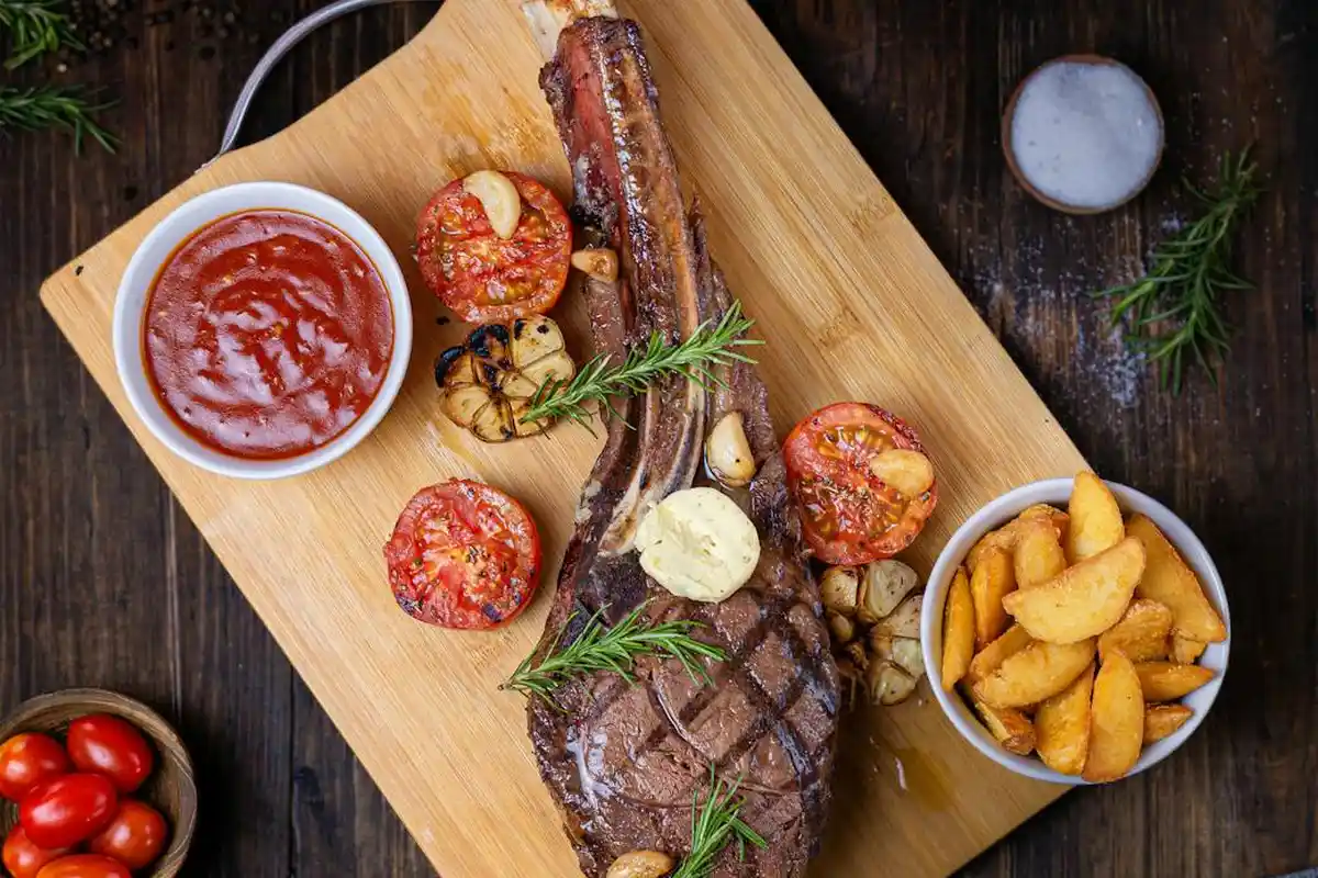 A wooden cutting board with a grilled steak, roasted cherry tomatoes, garlic, and potato wedges, garnished with rosemary.