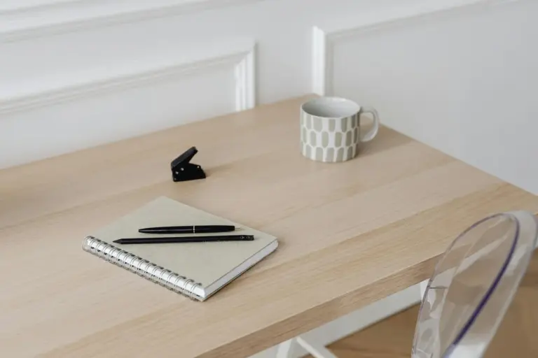 A light wooden desk with a notebook and pen, a small black stapler, a ceramic mug, and a transparent chair; a tidy workspace prepared for refinishing projects.