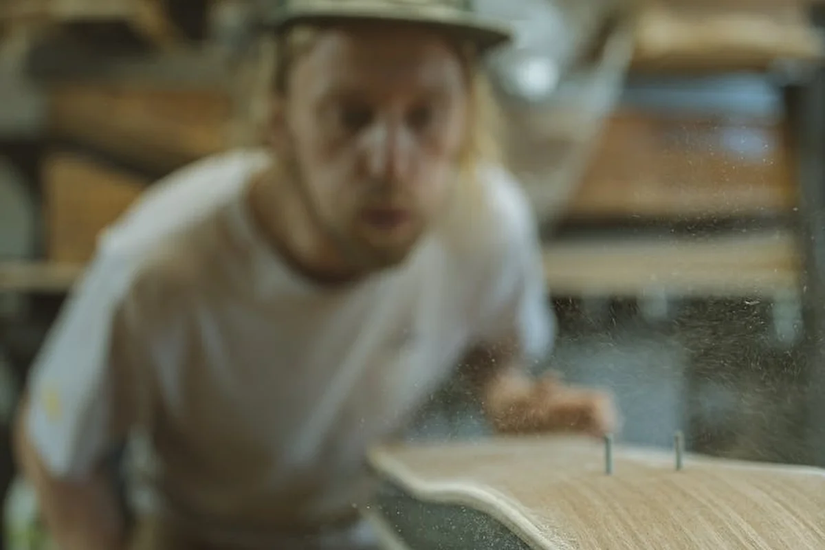 Blurry image of a person in a workshop inspecting a wooden board covered in dust, emphasizing wood dust exposure