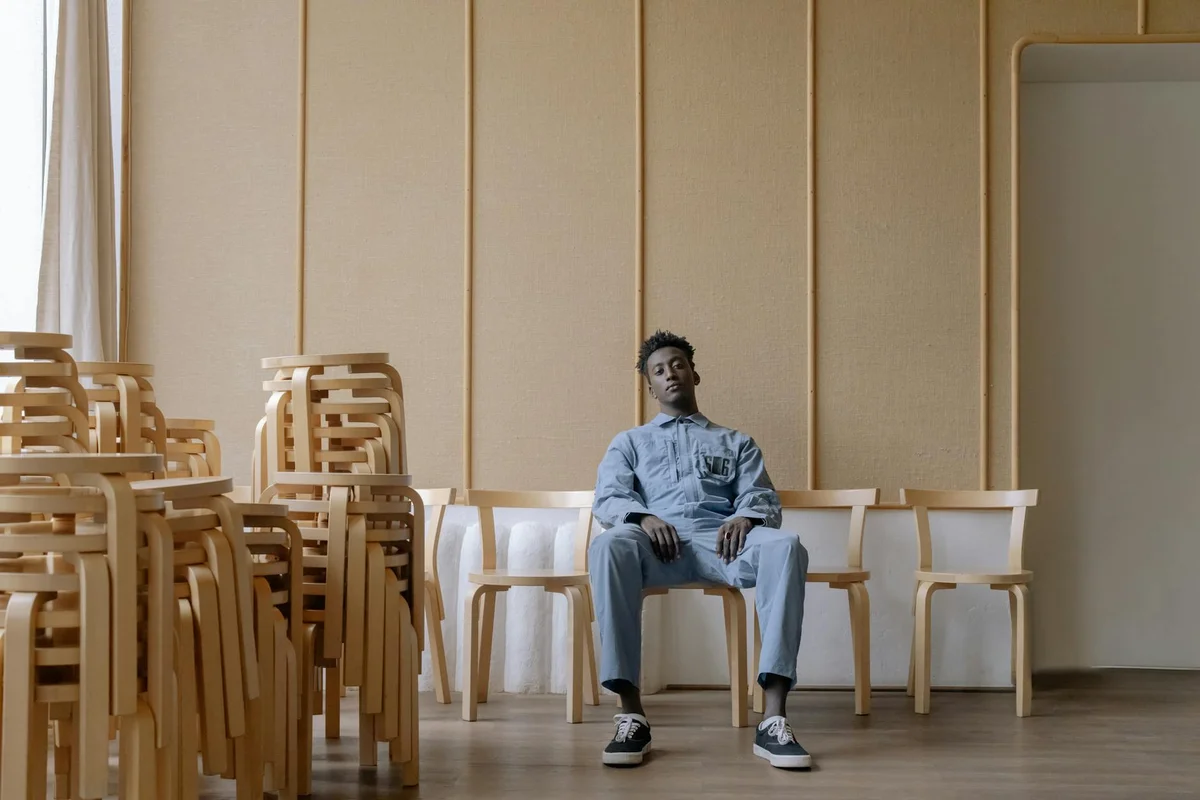 A person sits on a wooden chair in a bright, minimalist room with stacked wooden chairs lining the wall, suggesting a workspace for furniture finishing.