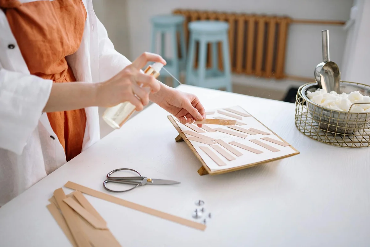 Close-up of a person applying glue to narrow wooden strips laid out on a white workspace; scissors, glue bottle, and other crafting tools nearby with a wicker basket and stools in the background.