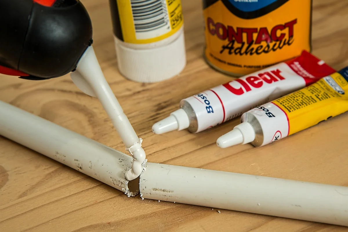 Close-up of a glue applicator applying adhesive to a joint on a wooden surface, with glue tubes and adhesive bottles visible in the background