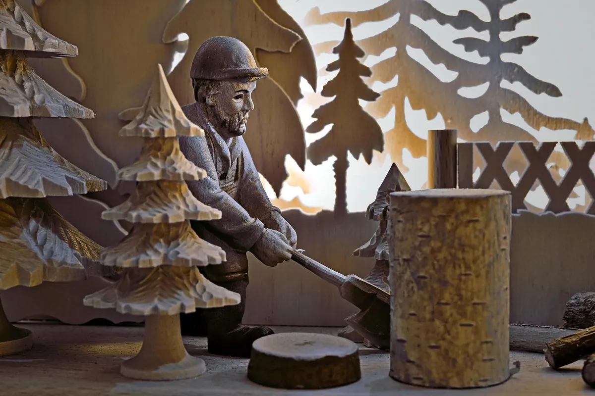Miniature woodworking scene: a lumberjack figurine wearing a helmet and using a tool on a wooden log, with carved trees and workshop backdrop.