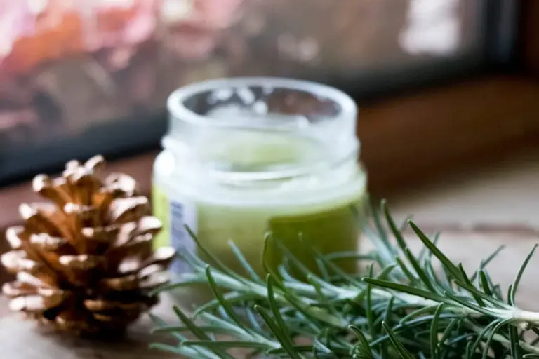 A small glass jar of greenish wood oil sits on a wooden surface, with a pinecone and a sprig of rosemary nearby.