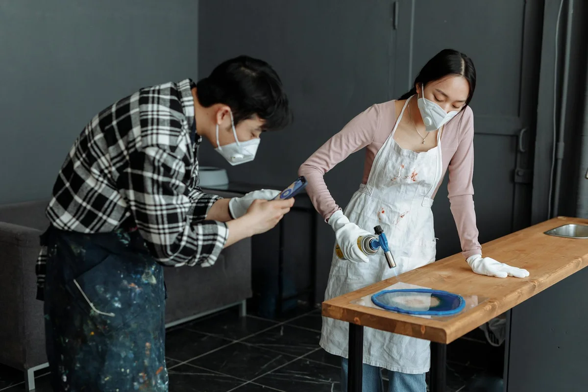 Two woodworkers wearing masks and aprons repair a wood table finish. One person leans over a workbench inspecting a tool, while the other stands nearby holding a smartphone, with a heat-safe workspace and protective gloves.