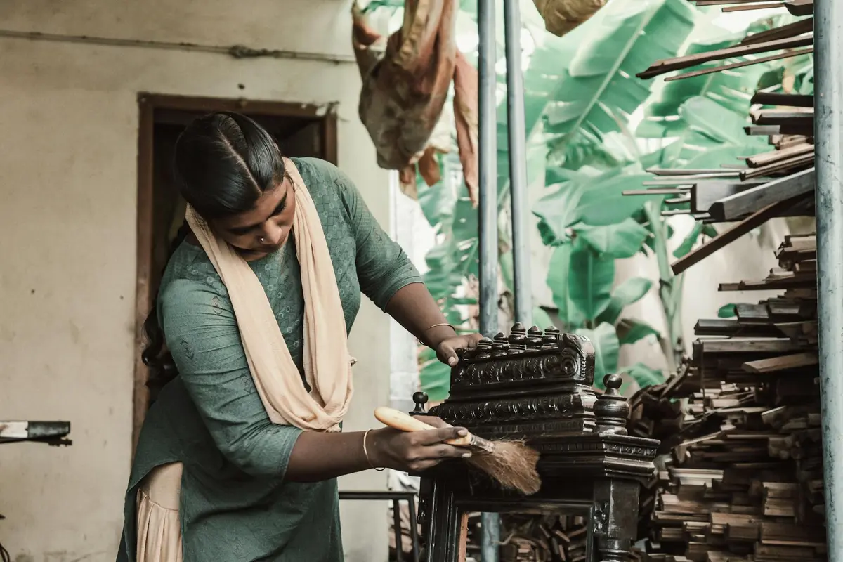 A woman wearing a green dress and scarf brushes a wooden piece in a workshop, with stacked wood and tools visible around.