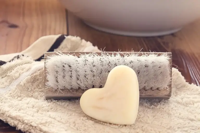 A brush with white bristles lying on a textured towel beside a heart-shaped bar of soap, suggesting manual cleaning of wood finishing tools.