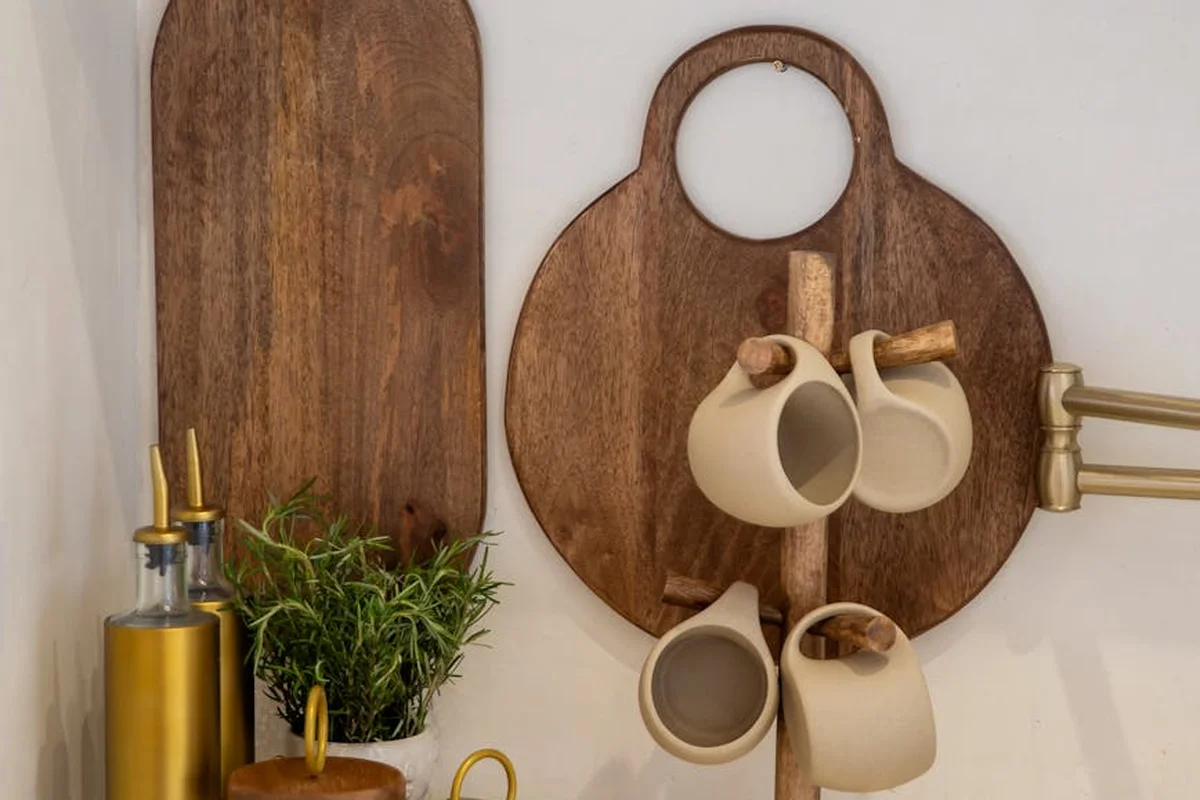 Wooden kitchen scene with two vertical cutting boards mounted on the wall and a circular mug rack holding several ceramic cups; a bottle of olive oil and a small potted herb sit on the counter.