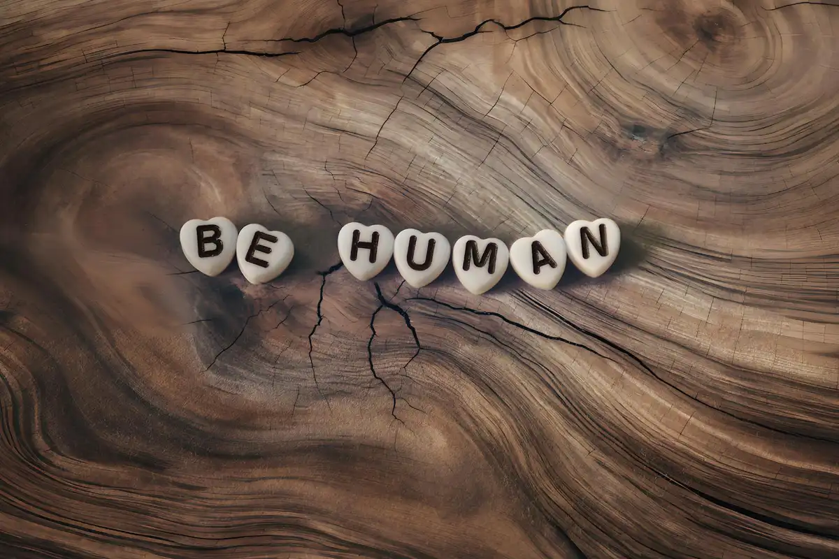 Close-up of a richly grained wooden surface with visible cracks; small beads spell out 'BE HUMAN' resting along the grain.