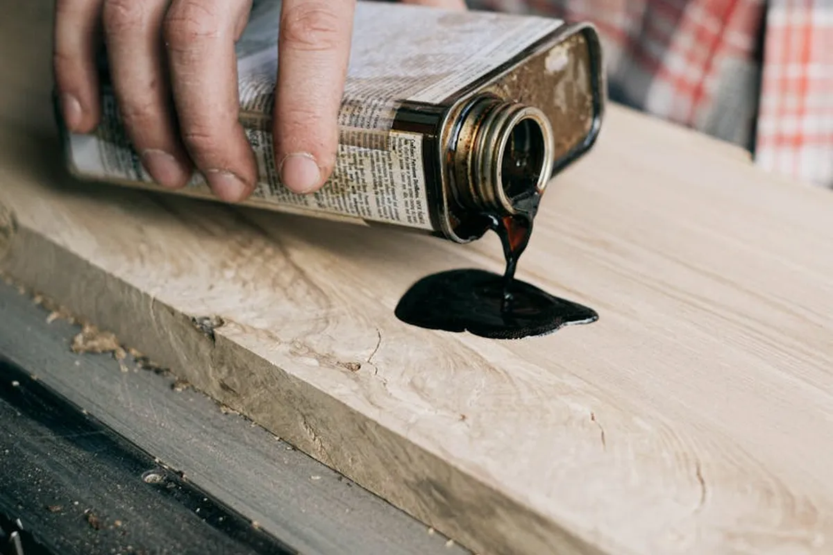 Close-up of a person pouring dark wood finish from a can onto a wooden workpiece, highlighting the need for ventilation in woodworking.