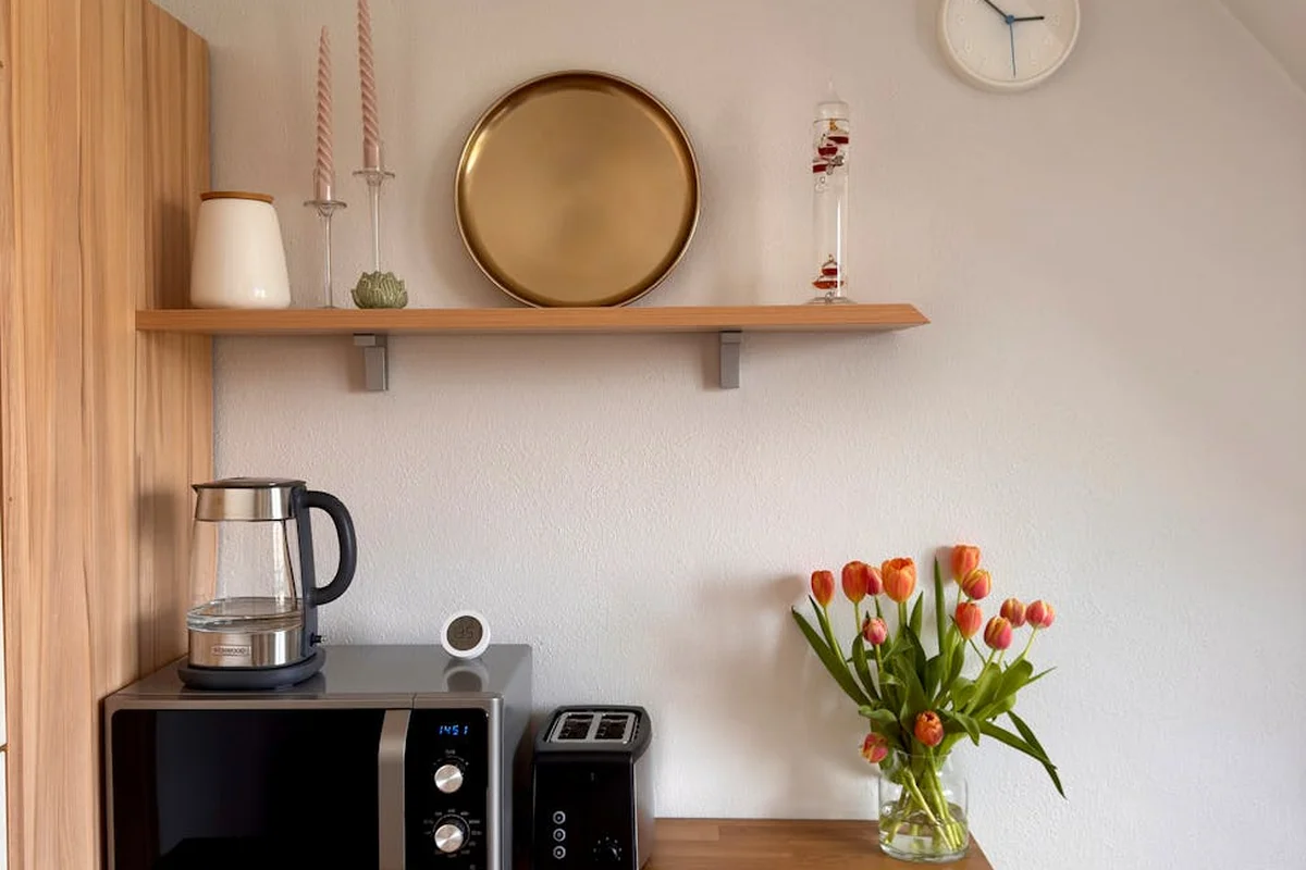 Bright kitchen scene with a microwave, kettle, and toaster on a wooden countertop; a vase of orange tulips and a shelf with decorative items sit against a white wall.