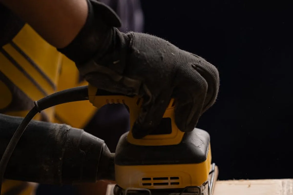 Close-up of gloved hands using a yellow handheld sander on a wooden surface, with dust and focus on the tool.