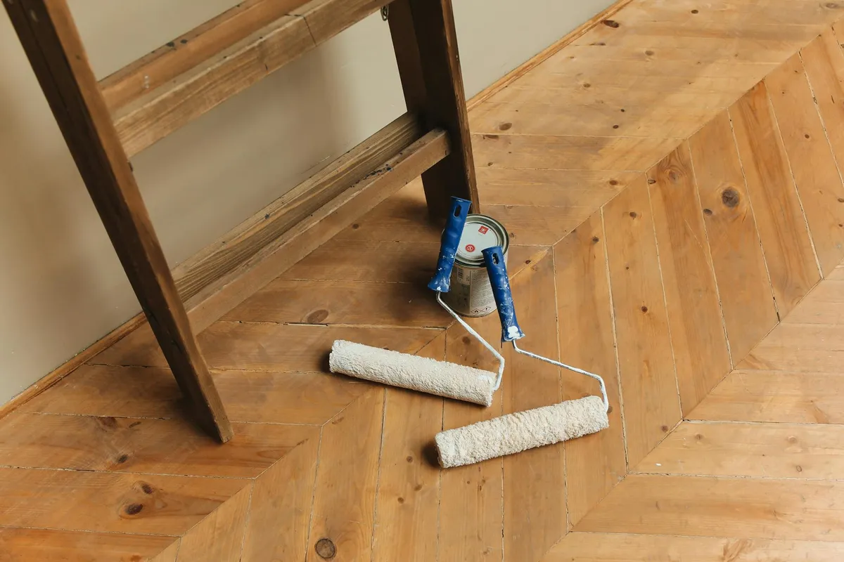 Two paint rollers and a small tool lie on a wooden floor next to a chair leg, suggesting wood finishing work in progress in a high-humidity environment.
