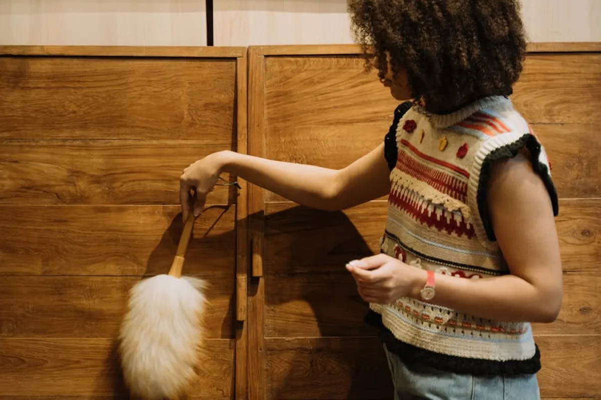Person with curly hair wearing a patterned sleeveless sweater dusting a wooden dresser with a fluffy brush
