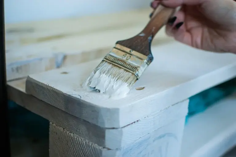 Close-up of a paintbrush applying a light coating to pale wood furniture.