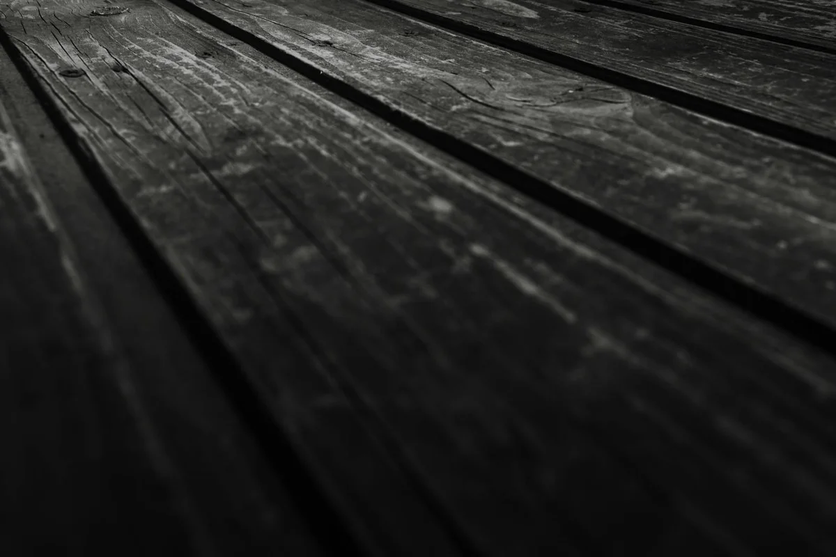Close-up view of dark, weathered wooden boards with visible grain and texture, set in a workshop environment.