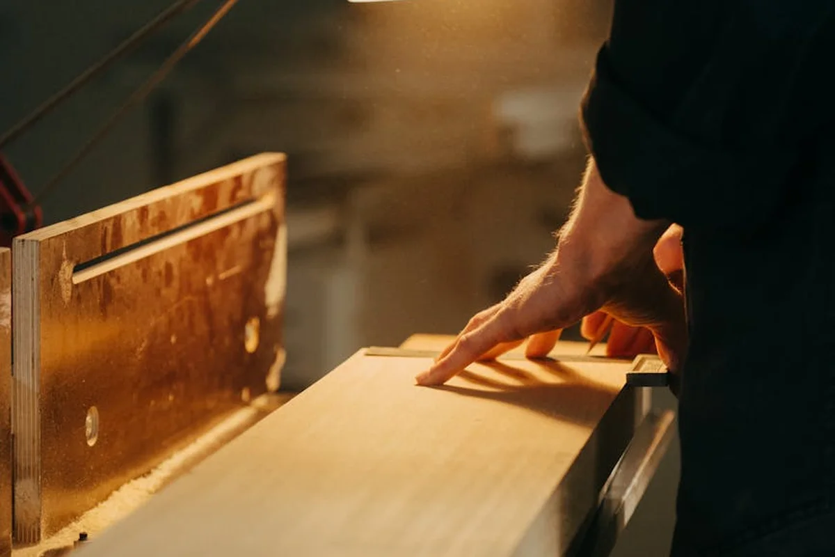Close-up of a woodworker guiding a wooden board through a table saw in a workshop.