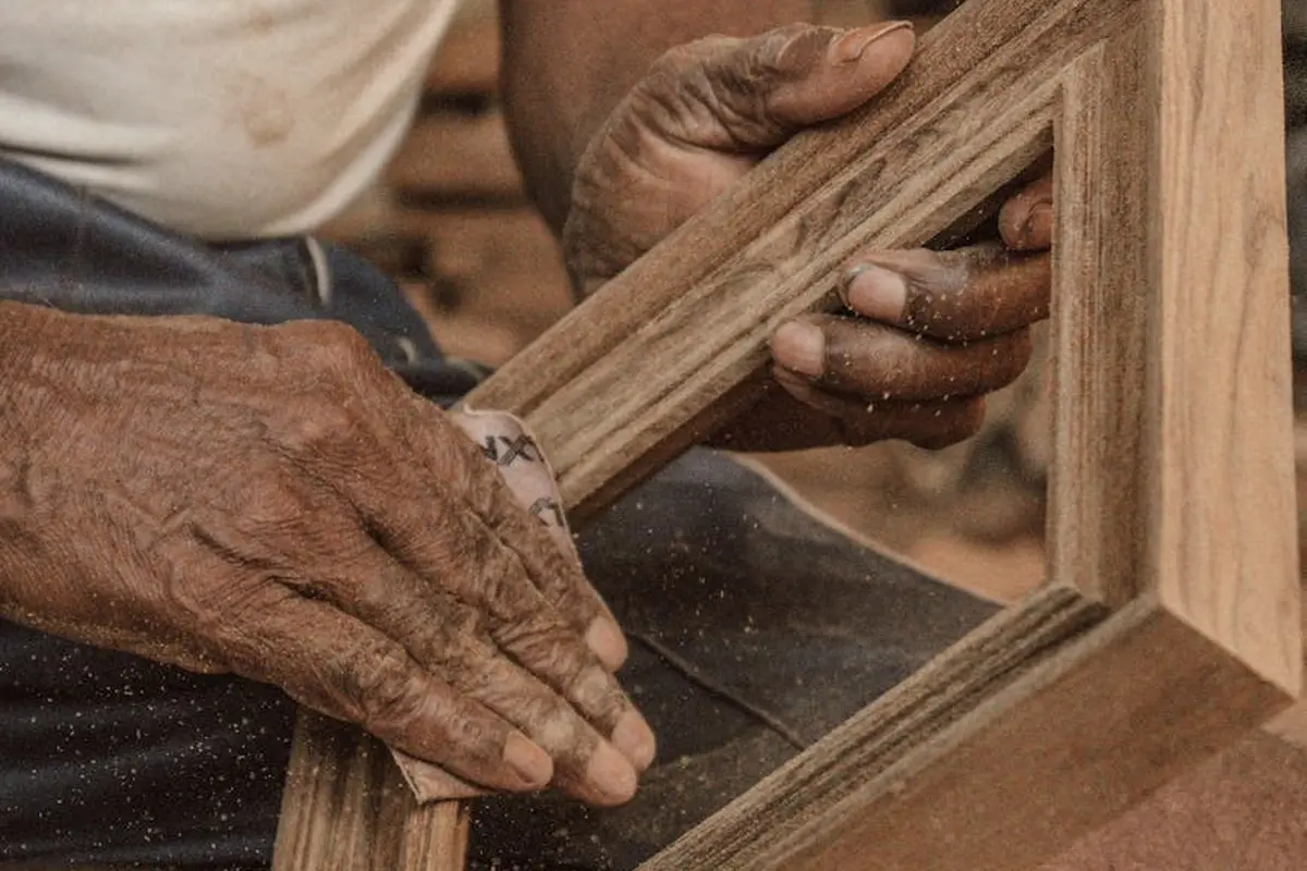 Close-up of a woodworker's hands sanding a wooden frame, with sawdust in the air and grain detail visible.