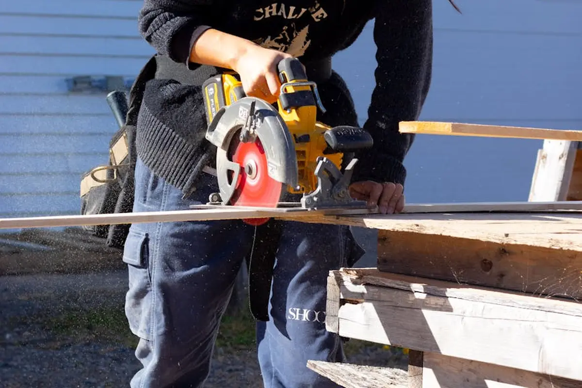 Person operating a yellow power circular saw to cut a wooden beam, working outdoors with wooden planks on a worktable or sawhorses.